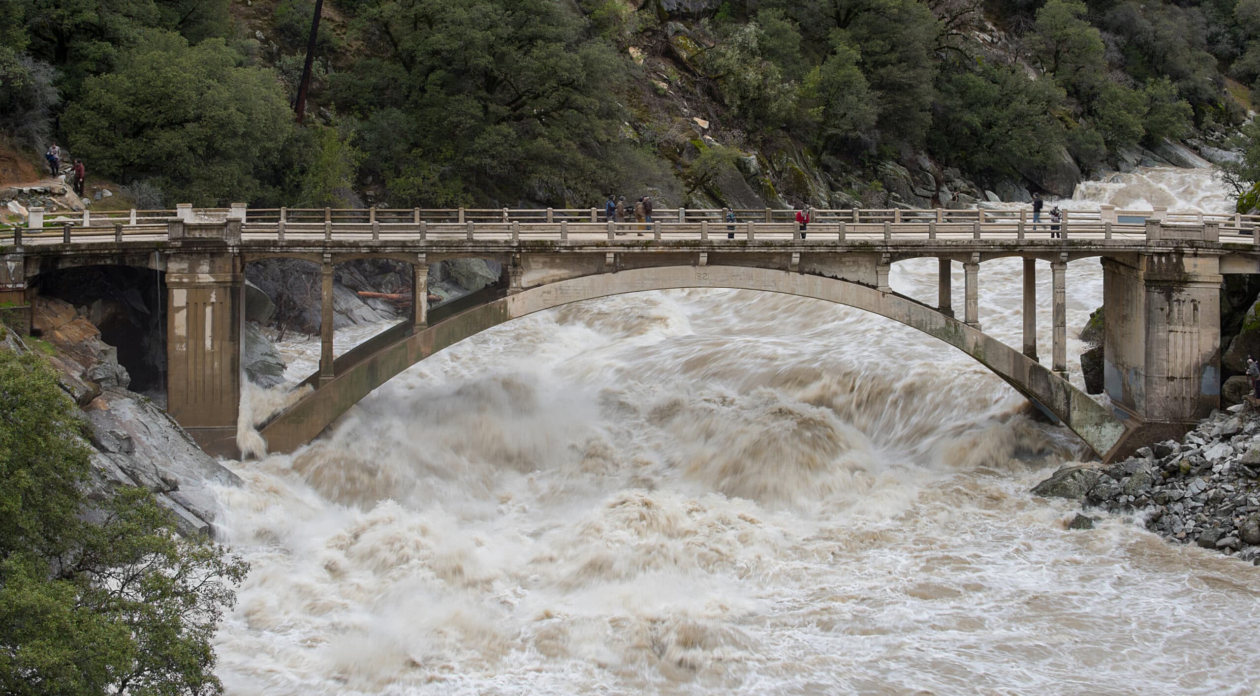 A photograph of a bridge suspended between two rocky hills. Underneath, foamy, brown water rages downstream. A family sits on the bridge watching the water. (via news.agu.org)